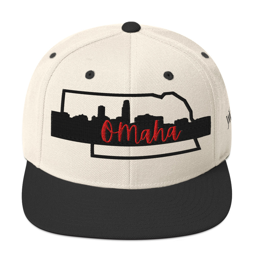 Black and beige snapback cap with the word Omaha embroidered in red against the silhouette of Nebraska and downtown Omaha.