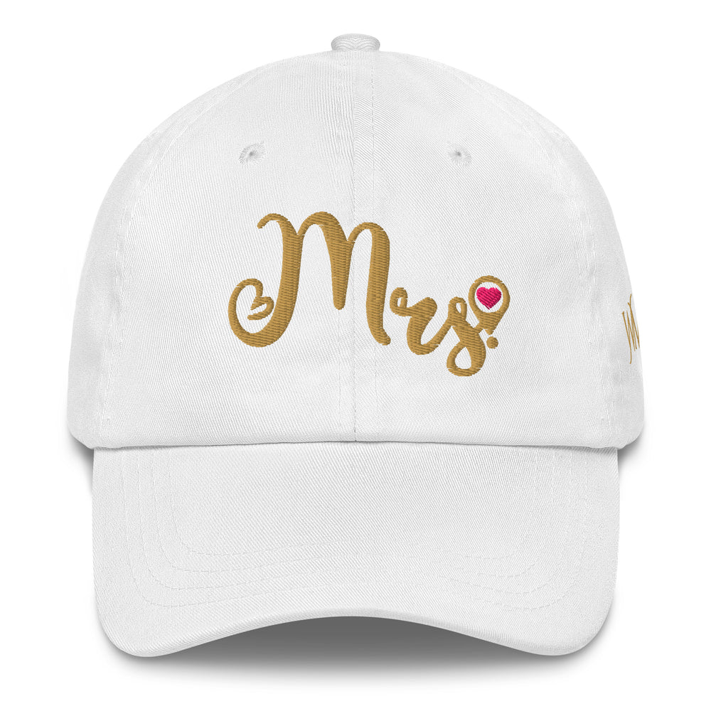 Celebrate the tradition of destination weddings with a white classic cap, with the word Mrs cursively embroidered in gold on the front. The tail of the S in Mrs has a location icon with a pink heart.