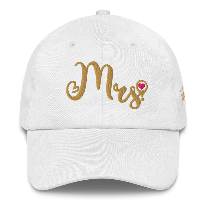 Celebrate the tradition of destination weddings with a white classic cap, with the word Mrs cursively embroidered in gold on the front. The tail of the S in Mrs has a location icon with a pink heart.