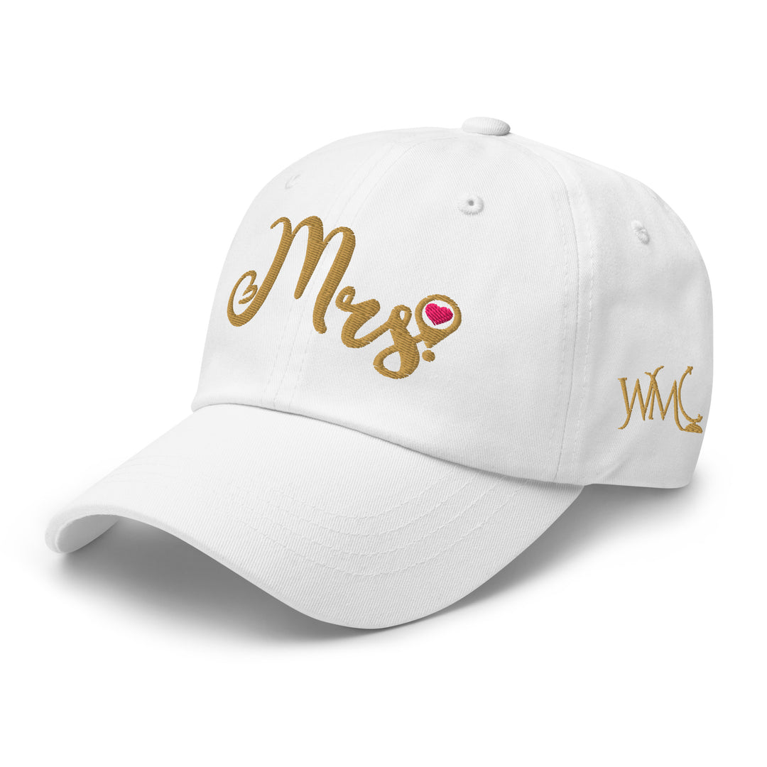 White classic cap with the word Mrs embroidered in gold in the front of the cap. The design is accompanied by a location icon with a pink circle in the middle.